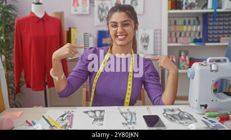 A smiling young hispanic woman wearing glasses and a measuring tape pointing at herself in a vibrant tailor shop. Stock Photo
