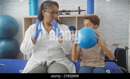 A woman physiotherapist instructs a young boy during a therapy session with a blue exercise ball in a rehab clinic. Stock Photo