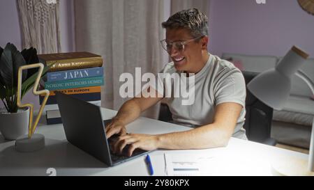 Young man smiling while using a laptop at home with a stack of interior design books, table lamp, and documents on a white desk in the living room Stock Photo