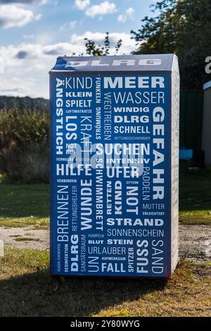 A blue changing cabin at a bathing spot on the Achterwasser lagoon displays a word cloud of water-safety terms – „danger", „sea", „search", „help", „lifeguard", „beach" – echoing the work of the DLRG (Deutsche Lebens-Rettungs-Gesellschaft), Germany's national life-saving association. Netzelkower Weg, Lütow, Gnitz peninsula, Usedom. Mecklenburg-Western Pomerania, Germany. Stock Photo