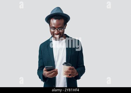 Handsome young African man using smart phone and holding disposable cup while standing against grey background Stock Photo