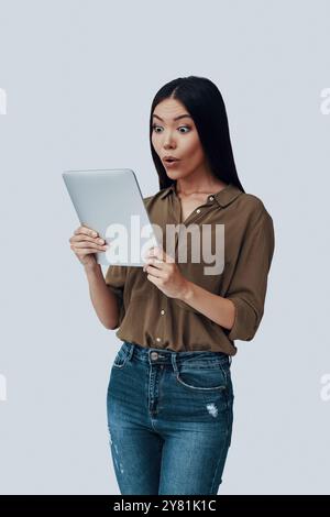 Wow! Surprised young Asian woman using digital tablet and making a face while standing against grey background Stock Photo