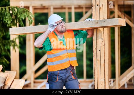 Carpenter constructing wooden-framed house near the woods. Bearded man with spectacles holds large beam on his shoulder and is dressed in work clothes and helmet. Modern construction concept. Stock Photo