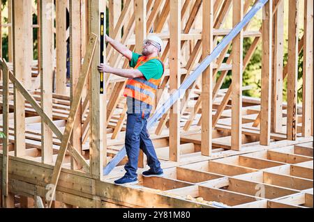 Carpenter constructing two-storey wooden frame house. Man inspects walls for levelness using spirit level, wearing protective overalls, helmet and vest. Concept of modern ecological construction. Stock Photo