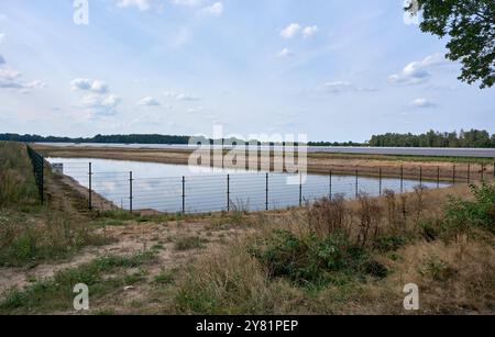 a large solar energy farm with pond surrounded by a fence featuring rows of photovoltaic panels in a rural area with trees and blue skies. Renewable e Stock Photo
