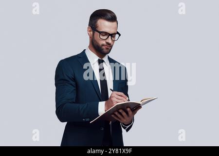 Making notes. Handsome young man writing something down while standing against grey background Stock Photo