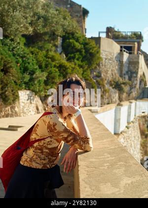 Smiling young woman leaning on a stone railing with a historical building in the background on a sunny day. Stock Photo