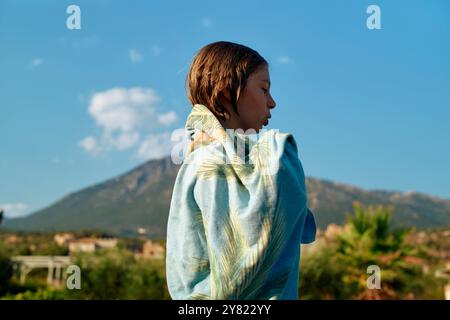 Young boy wrapped in a blue towel stands against a scenic backdrop of a distant mountain and clear sky. Stock Photo