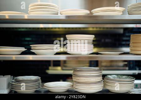 Stacks of various white ceramic plates on metal shelves in a commercial kitchen. Stock Photo