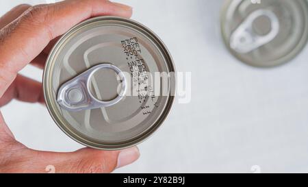 A close-up view of a hand holding a metal can with a pull-tab lid. The can's bottom shows expiration and production information. Another can is slight Stock Photo