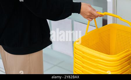 A person holding a bright yellow shopping basket in a retail environment. The basket is empty and stacked with other baskets in the background. The se Stock Photo