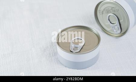 Two opened metal cans with pull-tab lids on a light textured surface. The cans have a simple design with a gray band around the bottom and a shiny met Stock Photo