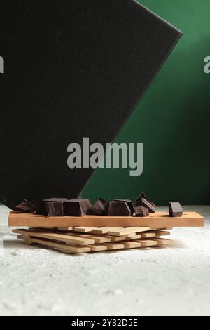 Chunks of chocolate arranged on a wooden board, diagonally tilted on a stone, with a white-green background, adding depth and dynamic visual appeal Stock Photo