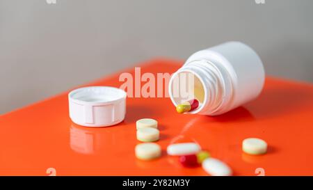 A white plastic pill bottle lying on its side with its cap off, spilling yellow and red capsules and white tablets onto an orange surface. Stock Photo
