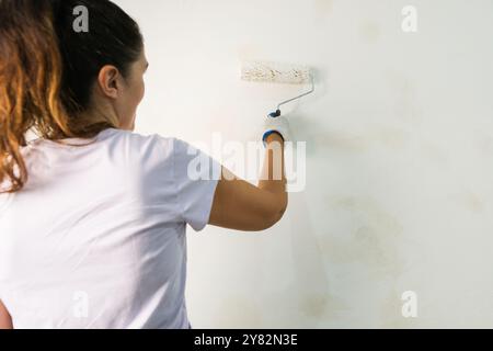 Back view of a young woman painting a wall with white paint using a roller Stock Photo