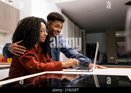 Smiling man embracing girlfriend using laptop standing in kitchen. Diverse carefree couple planing together with computer at home. Stock Photo