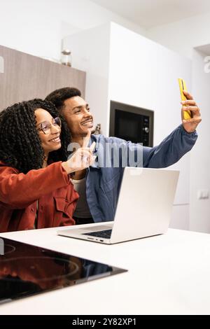 Smiling happy diverse couple taking a selfie while working online with laptop in their kitchen at home. Young adult man and woman taking a picture at Stock Photo