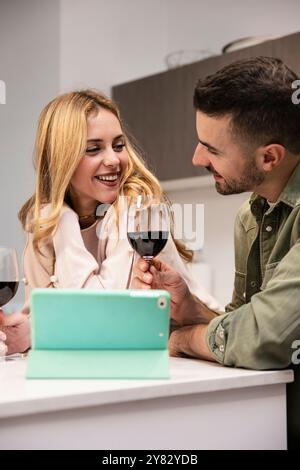 Young joyful couple enjoying wine at home using a tablet at home. Happy man and woman celebrating with drinks while using a touchpad in the kitchen. Stock Photo