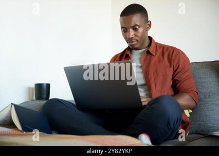 Focused young African man working remotely from home on a laptop while sitting on his living room sofa Stock Photo
