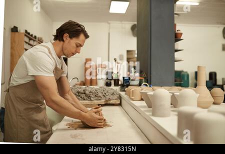 Young male potter wearing an apron shaping a wet piece of clay while working at a table in a ceramics studio Stock Photo