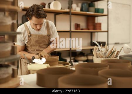 Young male ceramist wearing an apron shaping some clay into a bowl on a pottery wheel in a ceramics studio Stock Photo