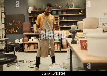 Young African potter wearing an apron using a mixer in a bucket of paint in a large ceramics manufacturing workshop Stock Photo