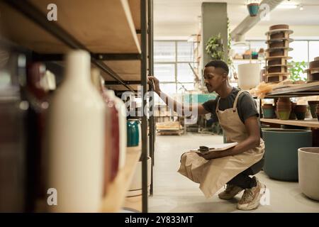 Young African pottery studio worker with a clipboard crouching in front of shelves of pots while taking inventory in a ceramics studio Stock Photo