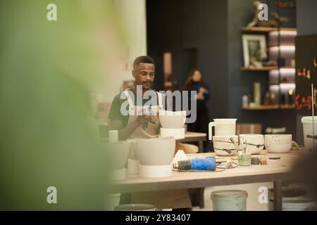 Young African potter cleaning a clay pot with a cloth while working in a large ceramics manufacturing studio Stock Photo