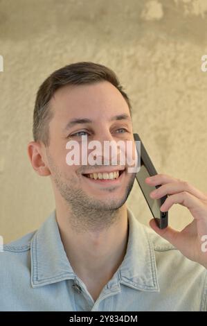 Close-up portrait of a guy with dark hair and stubble smiling while holding a phone near his face looking to the side Stock Photo