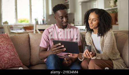 Smiling young multiracial couple using a digital tablet while sitting together on their living room sofa at home Stock Photo
