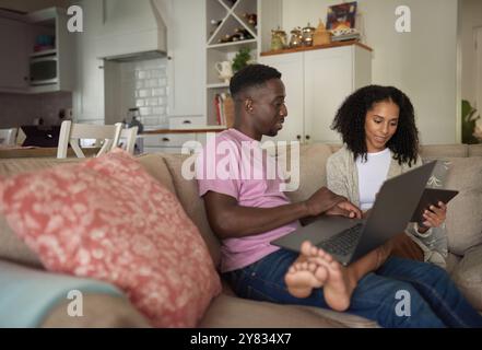 Smiling young multiracial couple working on a laptop and digital tablet while relaxing together on their living room sofa Stock Photo