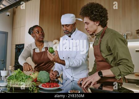 Mature chef in white uniform holding lime while demonstrating all types of ingredients to his biracial interns Stock Photo