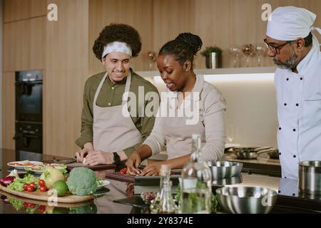 Young male sous chef helping African American female cook with cutting tomatoes and making caprese while chef in white uniform checking process Stock Photo