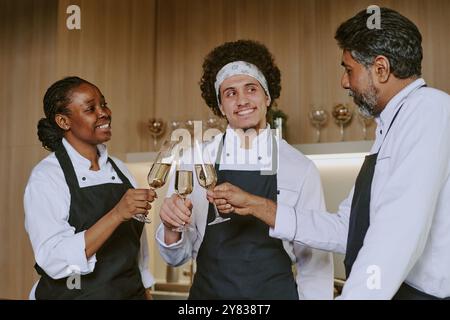 Multiracial cooking team clinking glasses of champagne while standing in center of spacious kitchen Stock Photo