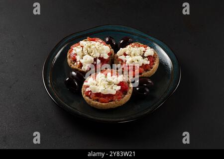Cretan dakos consists of barley rusk topped with juicy tomatoes, feta cheese and olive oil closeup on dark background. Stock Photo