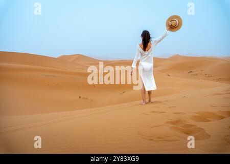 Young Brunette Woman in Camel Hat and White Dress Waving from Behind in Erg Chebbi Dunes Stock Photo