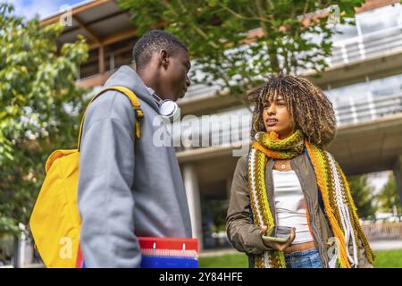 Low angle view photo of two diverse students talking and sharing mobile standing outside the university Stock Photo