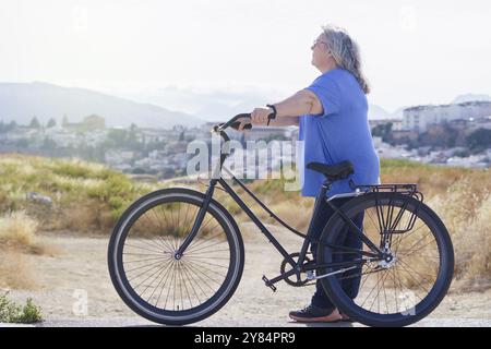 An overweight mature woman carries a black retro style bicycle, at the end of the path a small village and mountains with clouds in the background can Stock Photo