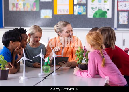 In school, female teacher with tablet, engaging diverse students with wind turbines and plants Stock Photo
