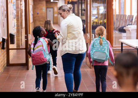 Walking in school hallway, female teacher guiding diverse students with backpacks to classroom Stock Photo