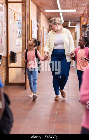Walking in school hallway, female teacher holding hands with two diverse students, smiling Stock Photo