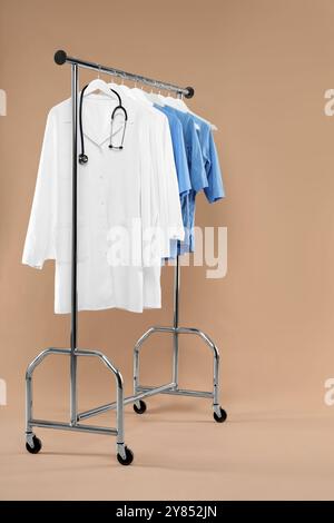 Different medical workers' uniforms and stethoscope on clothing rack against beige background Stock Photo