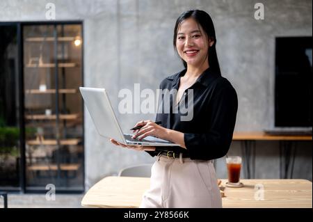 A confident, attractive Asian businesswoman is smiling at the camera while standing in her modern loft office with a laptop in her hand. Stock Photo