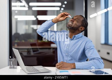Man sitting at office desk using eye drops to relieve discomfort from prolonged computer use. Wearing a blue shirt, he looks upward while applying eye drops. Stock Photo