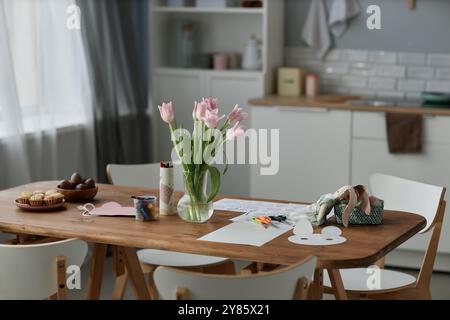 Wooden dining table adorned with Easter crafts and decorated eggs, bright tulips in glass vase adding freshness to the room, kitchen background with white cabinets visible Stock Photo
