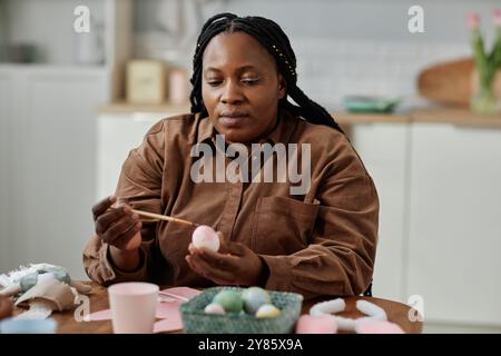 African American woman painting easter eggs in bright kitchen preparing for holiday celebrations creating festive atmosphere crafting artistic designs on eggs using brushes and colors Stock Photo