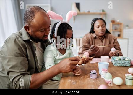 Family decorating Easter eggs together at home, enjoying creative time with various colors and brushes. Celebrating holiday tradition with parents and child wearing bunny ears headband Stock Photo