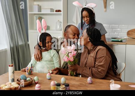Family enjoying Easter together by painting eggs and wearing bunny ears in cozy kitchen setting. Parents being affectionate while children observe with curiosity and delight Stock Photo