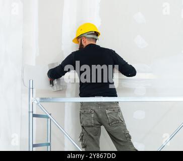 Construction worker wearing a hard hat and safety equipment plastering a wall using a trowel while standing on scaffolding Stock Photo