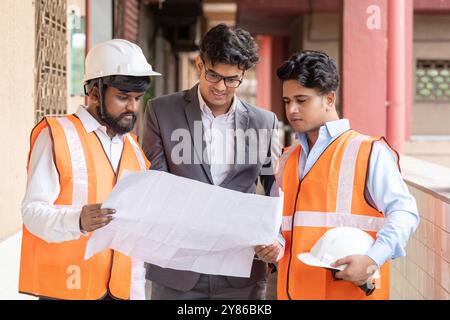 An architect, civil engineer, and interior designer closely examining a large set of building plans at a bustling construction site and discussing them. Stock Photo